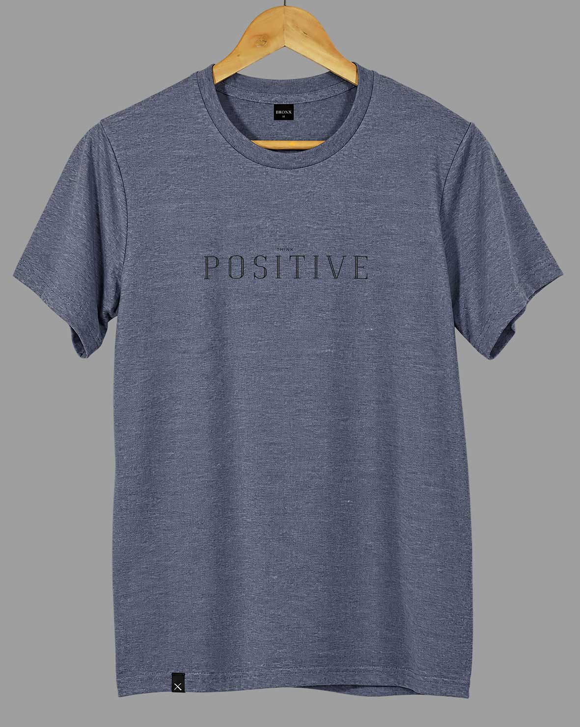 Polera Think Positive gris azulado BRONX colgada en percha de madera, mostrando su diseño frontal.