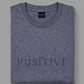 Polera Think Positive gris azulado de BRONX, doblada, con estampado frontal visible.