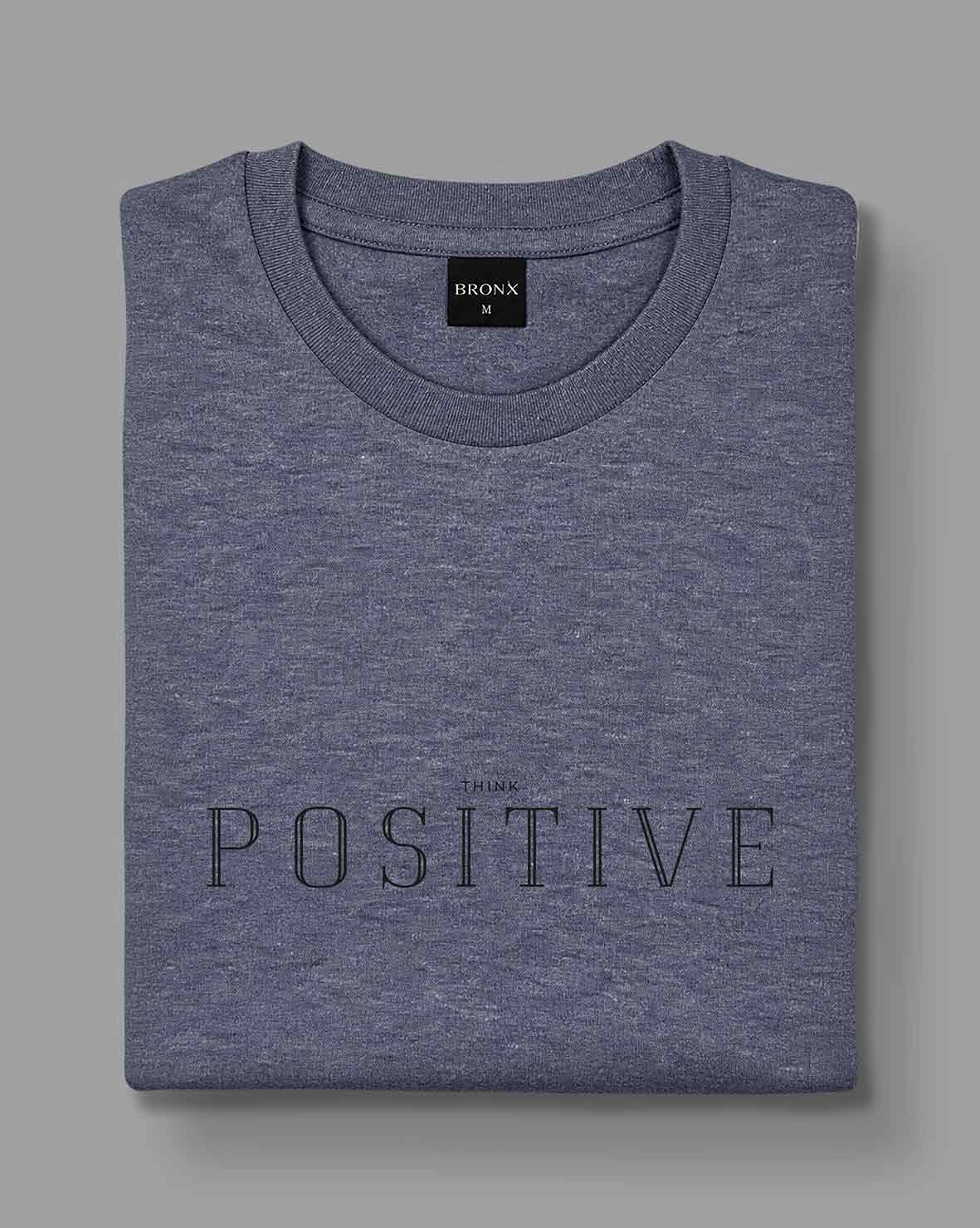 Polera Think Positive gris azulado de BRONX, doblada, con estampado frontal visible.